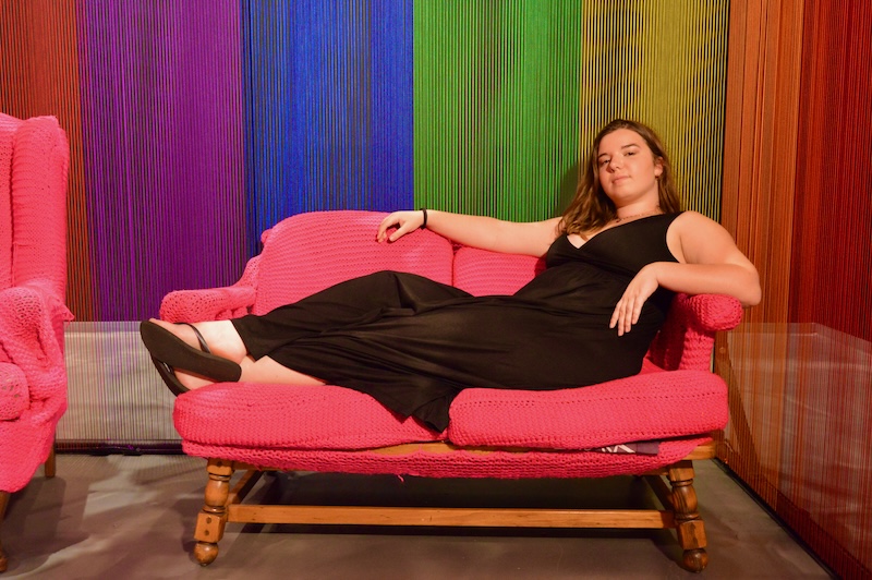 From the Wonder Art Gallery, this photo depicts a women laying on a pink couch with a rainbow yarn backdrop.